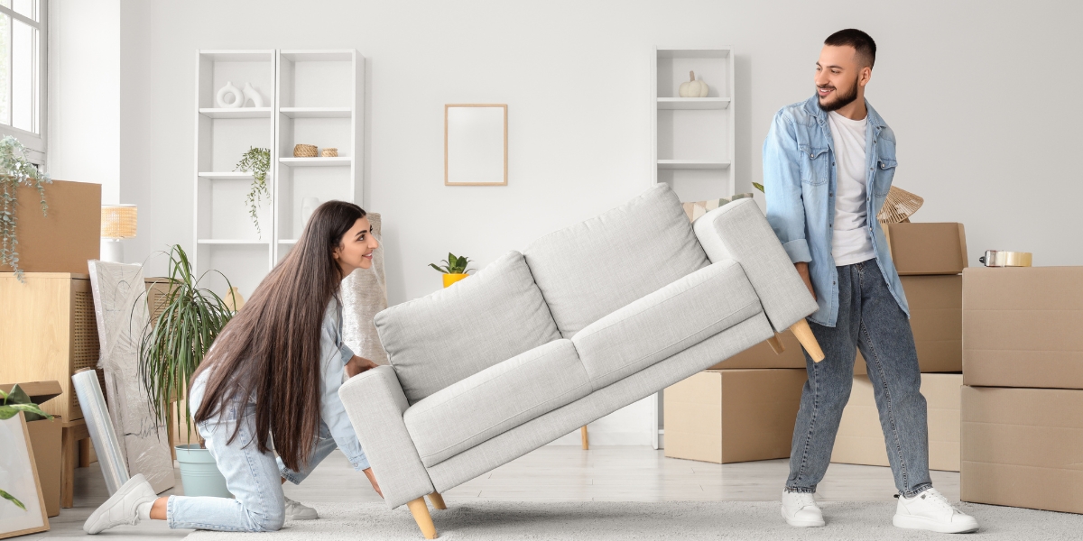 Starter home becomes a rental as young couple in denim moves gray couch surrounded by cardboard boxes in bright living room